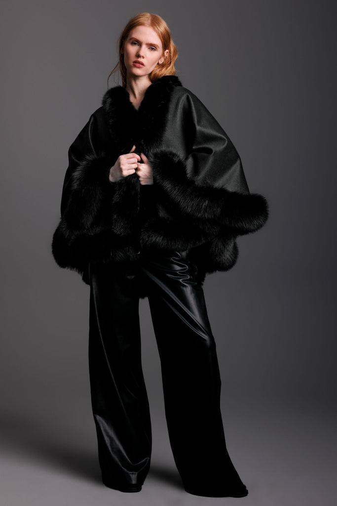 Woman wearing a black fur-trimmed cashmere cape in petite size and black leather pants 