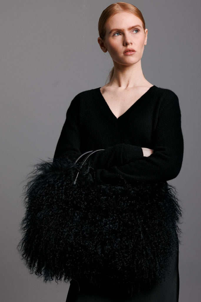 Woman holding a black mongolian lamb handbag with leather handles clutch against a gray background and wearing black silk dress