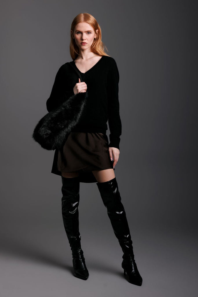 Woman wearing a black outfit with knee-high boots holding a fur shearling bag against a gray background