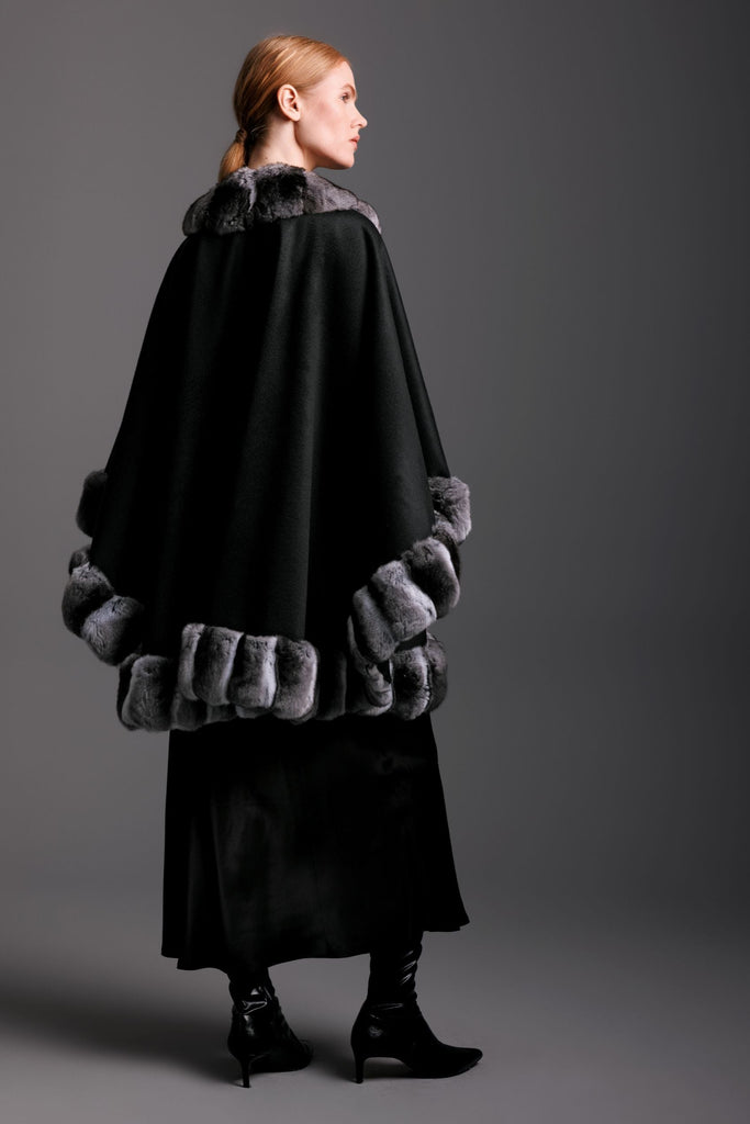 Woman wearing black cashmere cape with chinchilla fur trim all around and black slip dress and boots with a detail view of the backside.