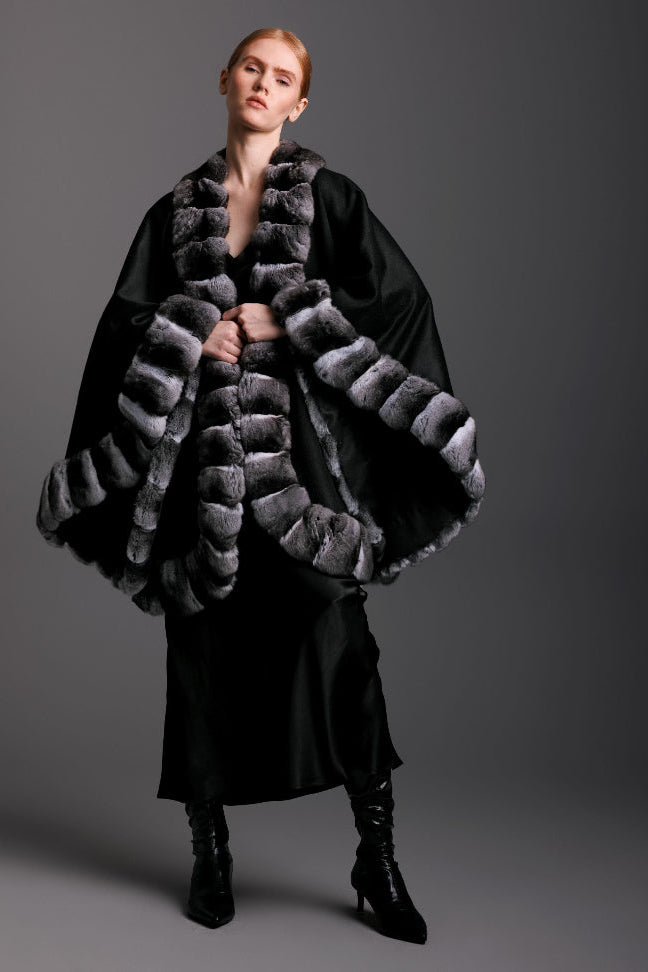 Woman wearing black cashmere cape with chinchilla fur trim all around and black slip dress and boots